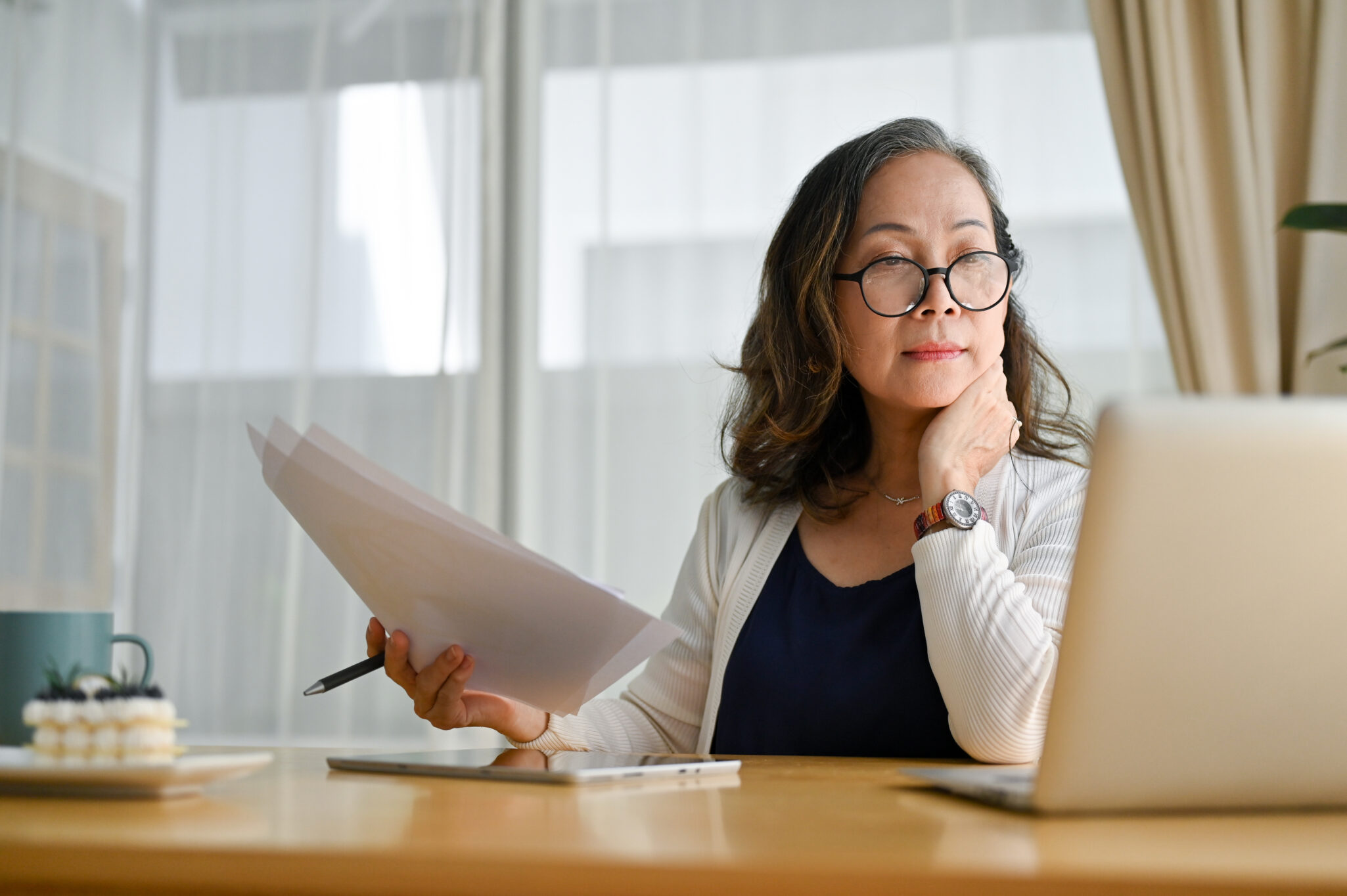 An older woman looks away from the paperwork she is holding and looks at the laptop in front of her