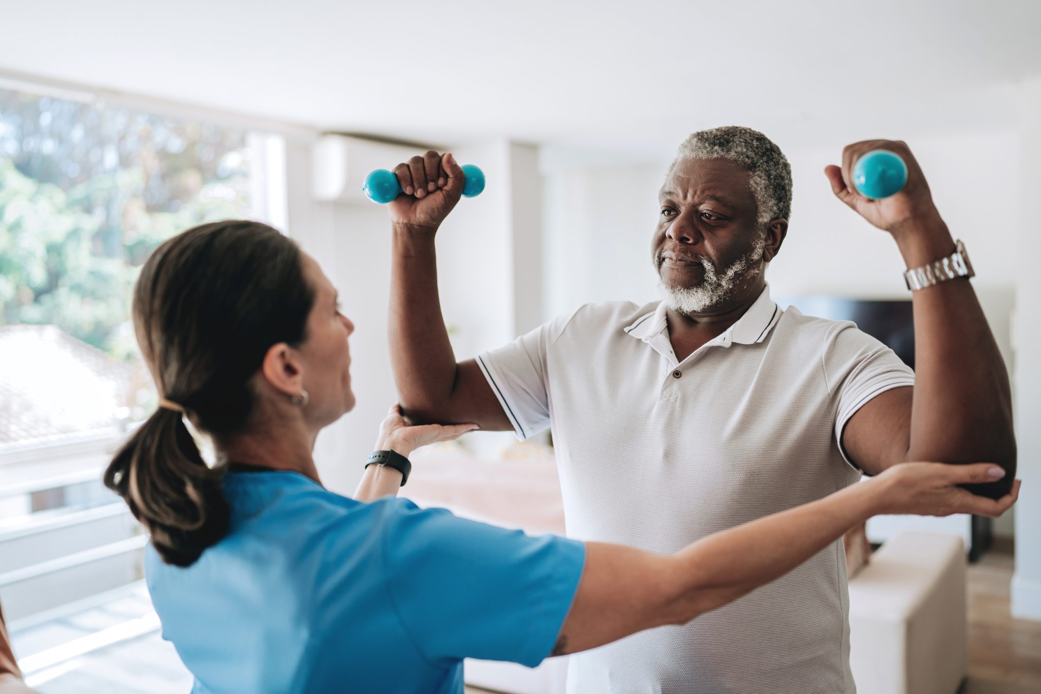 Senior man holding up dumbbells while female nurse is assisting him